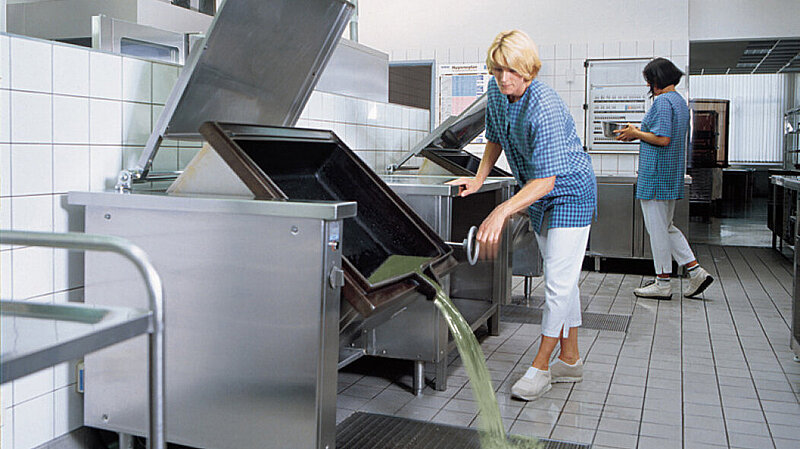 A large cooking pot is emptied into a floor tray in a commercial kitchen. A large cooking pot is emptied into a floor tray in a commercial kitchen.
